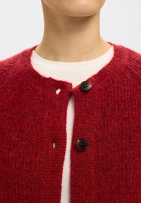 Raudonas megztas kardiganas su apvalia iškirpte, dviem juodais mygtukais, tekstūruotu raštu ir maža skylute netoli mygtukų siūlės.