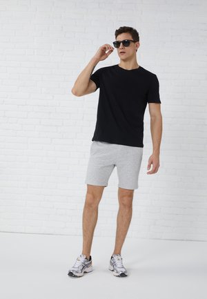 Uomo con maglietta nera, pantaloncini grigio chiaro, sneaker bianche e occhiali da sole che si aggiusta gli occhiali davanti a un muro di mattoni bianco.