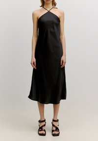 Zwarte satijnen slipdress met dunne kruislings strapjes, midi-lengte en een gladde textuur. Gecombineerd met bruine strap-sandalen.