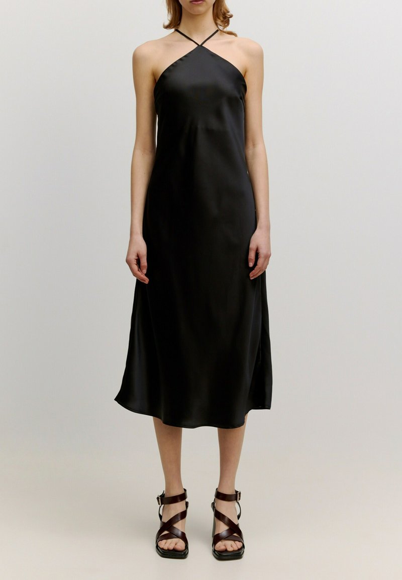 Zwarte satijnen slipdress met dunne kruislings strapjes, midi-lengte en een gladde textuur. Gecombineerd met bruine strap-sandalen.