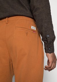 Camisa castanha com botão e tecido texturizado, combinada com calças laranjas com um bolso traseiro e uma etiqueta da Levi's. Tecido com textura suave.