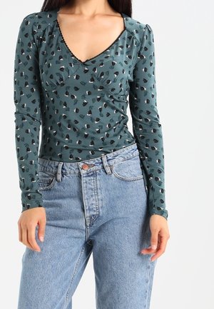 Woman wearing green long-sleeve top with small black and white floral pattern tucked into high-waisted light blue jeans.