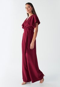 Burgundijski kombinezon iz lahkega blaga, z ovijalnim dizajnom s kratkimi rokavi, pasom za zavezovanje in širokimi hlačnicami.