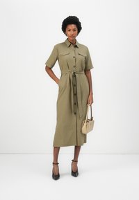 Ελαιόχρωμο φόρεμα με κουμπιά και κοντά μανίκια, δύο τσέπες στο στήθος, στενή μέση με ζώνη; συνδυασμένο με μαύρες γόβες και μπεζ τσάντα.