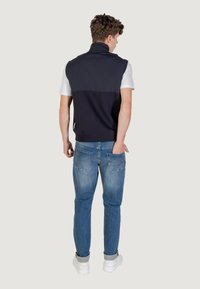 Gilet senza maniche di colore blu navy scuro sopra una camicia bianca, abbinato a jeans blu chiaro e scarpe da ginnastica bianche. Texture liscia, design semplice, senza motivi visibili.