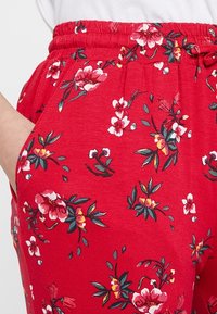 Röda blommiga byxor med elastisk midja, sidofickor och ett mjukt tyg med ett livligt blommönster i nyanser av rosa och grönt.