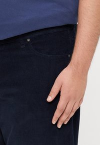 Navy corduroy pants with vertical ribbing, two pockets, and metal button accents. Hand resting on the thigh, displaying soft, textured fabric.