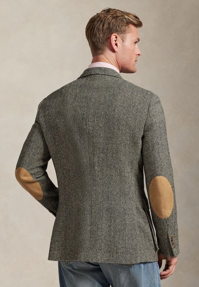 Homme portant une veste gris chevron avec des coudières couleur fauve et un jean bleu, vu de profil sur un fond uni.