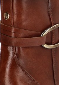 Botín de cuero marrón que presenta un diseño curvado, textura suave y un detalle decorativo de hebilla y correa de metal.