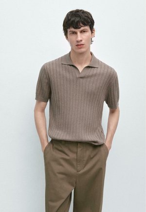 Jeune homme aux cheveux foncés portant un polo à manches courtes en maille côtelée marron et un pantalon marron, debout avec les mains dans les poches.