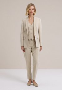 Bež odijelo po mjeri s blejzerom na jedno dugme, prslukom u istoj boji i hlačama ravnih nogavica. Glatki materijal, minimalistički dizajn i diskretni gumbi.