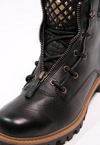 Botte en cuir noir avec fermeture éclair latérale partiellement ouverte, lacets ronds, œillets métalliques, languette cloutée et semelle marron robuste.