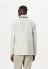 Light beige jacket with a contrasting brown collar, straight cut, long sleeves, and smooth texture, worn over light gray trousers.