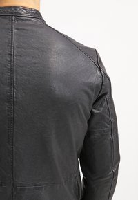 Vista posteriore di una persona che indossa una giacca di pelle nera con dettagli delle cuciture visibili sulle spalle e sulla schiena, su uno sfondo bianco.