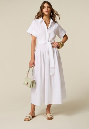 Vrouw draagt een witte midi-jurk met ceintuur, houdt een lichtgroene handtas vast, geaccentueerd met gouden armbanden, oorringen en sandalen.