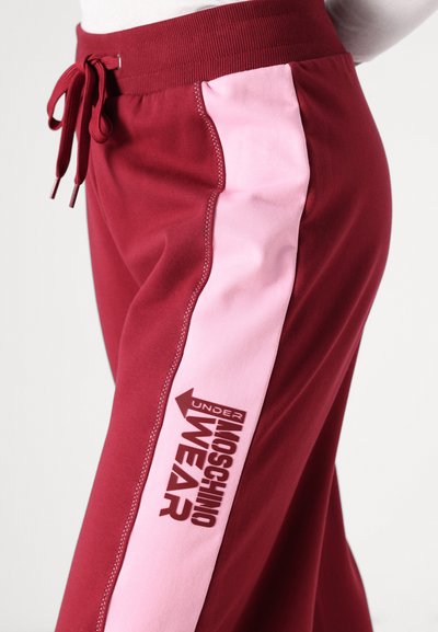 Bordo spalvos sportinės kelnės su rožinėmis šoninėmis juostomis; turi raištinį juosmenį ir spausdintą logotipą ant apatinės kojos. Minkšta medžiaga su lygiu paviršiumi.