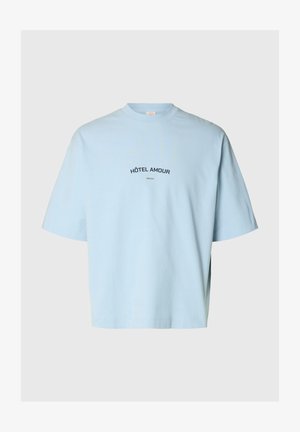 Lyseblå T-shirt med korte ærmer med teksten "HÔTEL AMOUR" og en lille "Selected" tekst centreret på brystet, ensfarvet baggrund.
