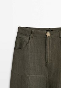 Jupe taille haute couleur olive avec boutons à l'avant, passants pour ceinture, et poches avant courbées, fabriquée en tissu texturé.