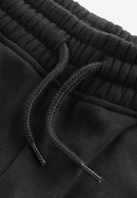 Schwarze Sport-Jogginghosen mit einem elastischen Bund und geflochtenen Kordeln mit Metallenden, die eine glatte Stofftextur und eine einheitliche Farbe aufweisen.