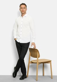 Weißes Langarmhemd mit Knopfverschluss, kombiniert mit schwarzen Hosen und schwarzen Schuhen. Hellholzgünstiger Stuhl neben dem Model. Neutrale Hintergrund.