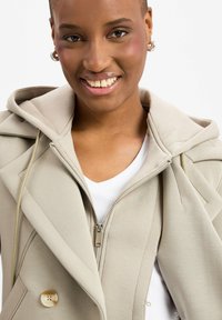 Helles beige Jacket mit Kapuze, doppelreihigem Design, Reißverschluss und sichtbarem Knopf. Texturiertes Material kontrastiert mit dem darunter liegenden weißen Hemd.