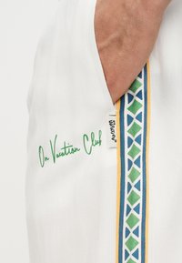 Detailný záber ruky v vrecku bieleho odevu s nápisom "On Vacation Club" v zelenej farbe a modrým, zeleným a žltým geometrickým vzorovaným lemom.