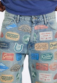 Dżinsy z denimu z wieloma kolorowymi naszywkami i logo Guess Originals w różnych kształtach i rozmiarach na przednich udach.