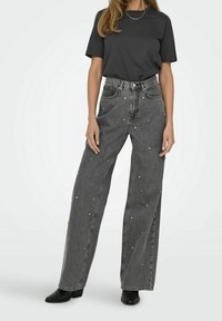 Jeans de pierna ancha en denim negro con adornos de cuentas plateadas, cintura alta, bolsillos clásicos y una textura ligeramente desgastada. Combinados con una camiseta negra.