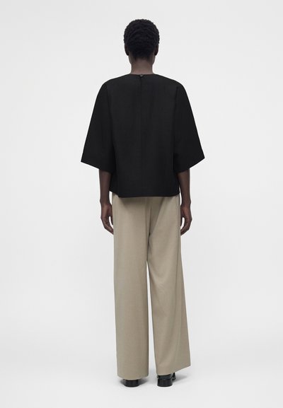 Άτομο με κοντά μαλλιά που φοράει μαύρο τοπ με κοντά μανίκια και μπεζ παντελόνι με φαρδιά πόδια, στέκεται με την πλάτη σε ένα απλό φόντο.
