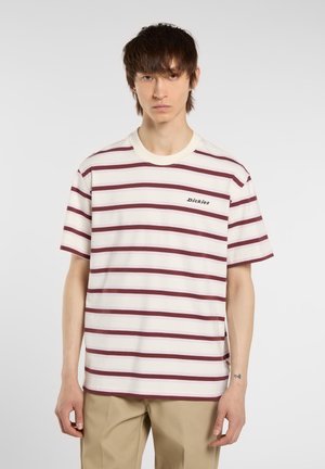 A person with medium-length hair wears a striped t-shirt featuring maroon and white colors and light pants, posing against a plain background.