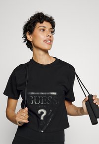 Crni pamučni majica s sjajnim crnim trokutastim grafikom s tekstom "GUESS" i upitnikom. Model drži užad za preskakanje.