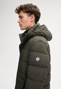 Giacca puffer verde con design trapuntato, cappuccio regolabile e un logo rotondo in evidenza sulla manica. Tessuto testurizzato con finitura opaca.