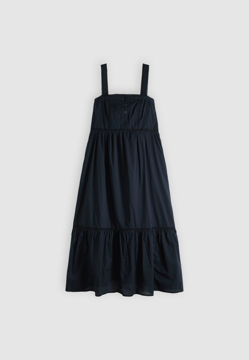Robe longue noire sans manches avec encolure carrée, corsage boutonné devant, volants froncés et larges bretelles sur un fond clair.