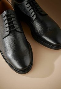 Sorte læder dress sko med en struktureret overflade, rund tå og seks snørrehuller, sat mod en neutral baggrund.