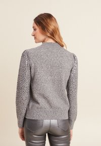 Grauer Pullover mit strukturiertem Strickkörper, gepufften Ärmeln, die mit Pailletten verziert sind, und geripptem Saum. Kombiniert mit glänzenden, metallischen Leggings.