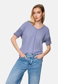 Helllila Kurzarmoberteil mit Rundhalsausschnitt und umgeschlagenen Ärmeln, kombiniert mit ausgebleichten blauen Jeans. Weicher Stoff mit sanftem Glanz.