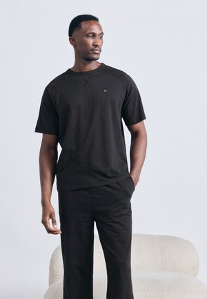 Mężczyzna w czarnej koszulce z krótkim rękawem i czarnych spodniach, stojący z jedną ręką w kieszeni, za nim jasnokolorowa, zakrzywiona sofa.