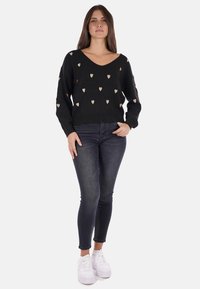 Maglione nero con scollo a V, caratterizzato da piccoli motivi a cuore beige. Abbinato a jeans scuri e sneakers bianche. Texture morbida, maniche lunghe.