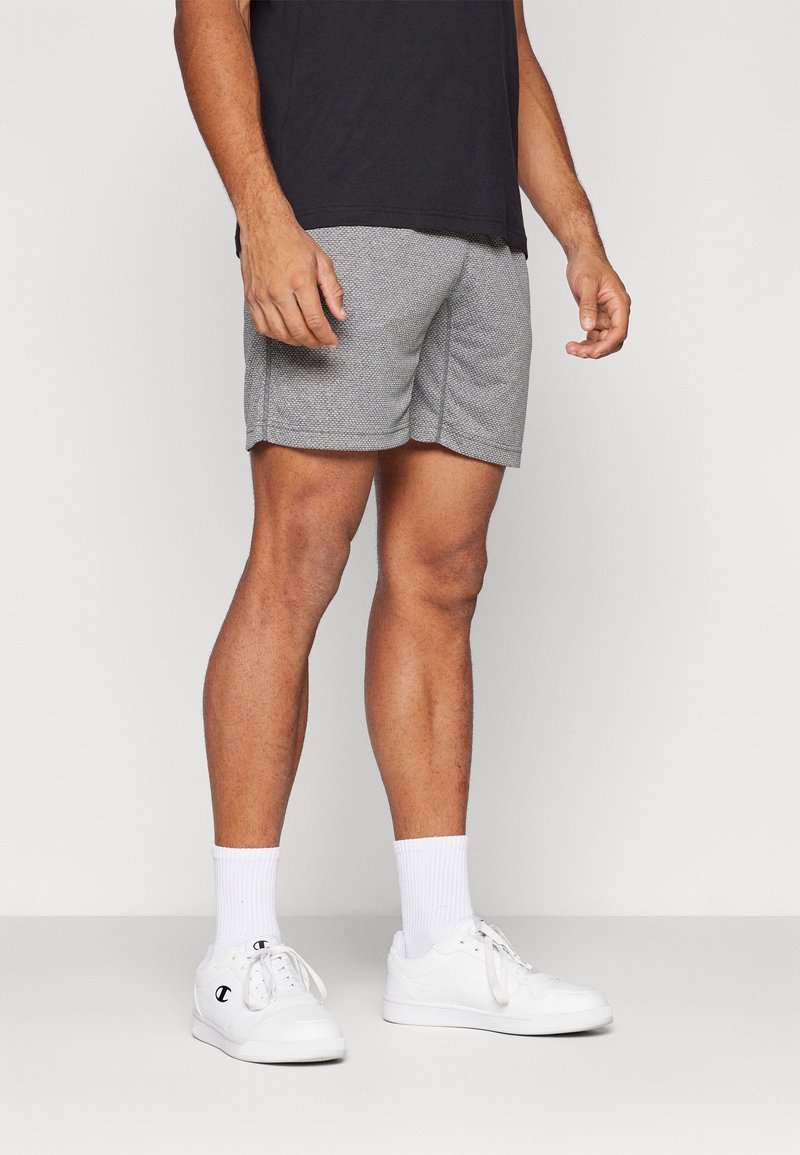 Homme portant un short gris texturé, des chaussettes blanches mi-hautes, des baskets blanches et un t-shirt noir, debout devant un fond uni.
