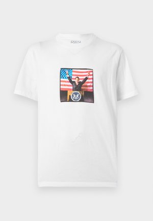 Camiseta blanca con un gráfico de un hombre con traje en un podio, con los brazos levantados, delante de una bandera de Estados Unidos.