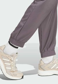 Pantalones deportivos de color púrpura claro con puños elásticos, combinados con zapatillas de correr beige con rayas blancas, malla texturizada y suelas en contraste.