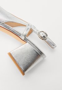 Silver metallic heeled sandal with a wide block heel, adjustable ankle strap, and a smooth texture. Features a circular buckle detail.