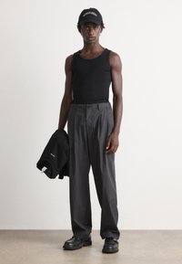 Slim person wearing black tank top, wide dark gray pants, black shoes, and black cap holding folded black sweatshirt in a plain room.