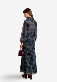 Maxikleid aus transparentem dunkelblauem Stoff mit schwarzem Blumenmuster, langen puffärmel, hohem Kragen und einem fließenden Schnitt. Kombiniert mit einer Mini-Handtasche.
