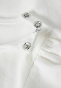 Weißes Baumwollkleidungsstück mit silbernen Druckknöpfen und gerafftem Stoffdetail, strukturierte Oberfläche und glatte Verarbeitung.