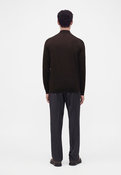 Homme debout de dos portant une chemise à manches longues sombre, un pantalon gris foncé et des chaussures foncées sur un fond clair uni.