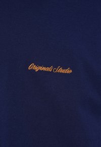 T-shirt blu navy a maniche corte, con scritto ricamato in arancione "Originals Studio" sul davanti. Materiale in cotone morbido, design minimalista.