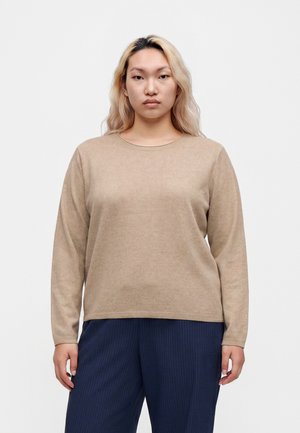 Giovane donna con lunghi capelli biondi che indossa un maglione beige e pantaloni a righe blu scuro, in piedi davanti a uno sfondo chiaro e uniforme.
