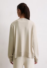 Donna con i capelli scuri raccolti in un ponytail, che indossa un maglione color crema e pantaloni abbinati, in piedi di fronte a uno sfondo bianco semplice.