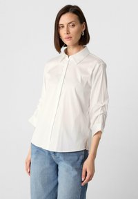 Weißes Hemd mit Knopfverschluss vorne, gerafften langen Ärmeln und seitlichen Schlitzen. Kombiniert mit hellblauen Jeans im minimalistischen Design.